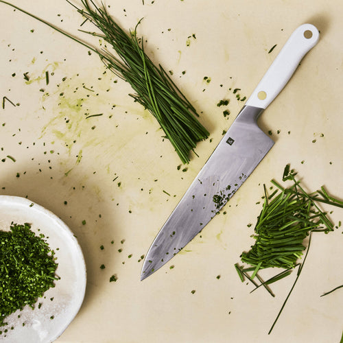Chef's knife resting on a counter beside finely chopped herbs and flaky salt