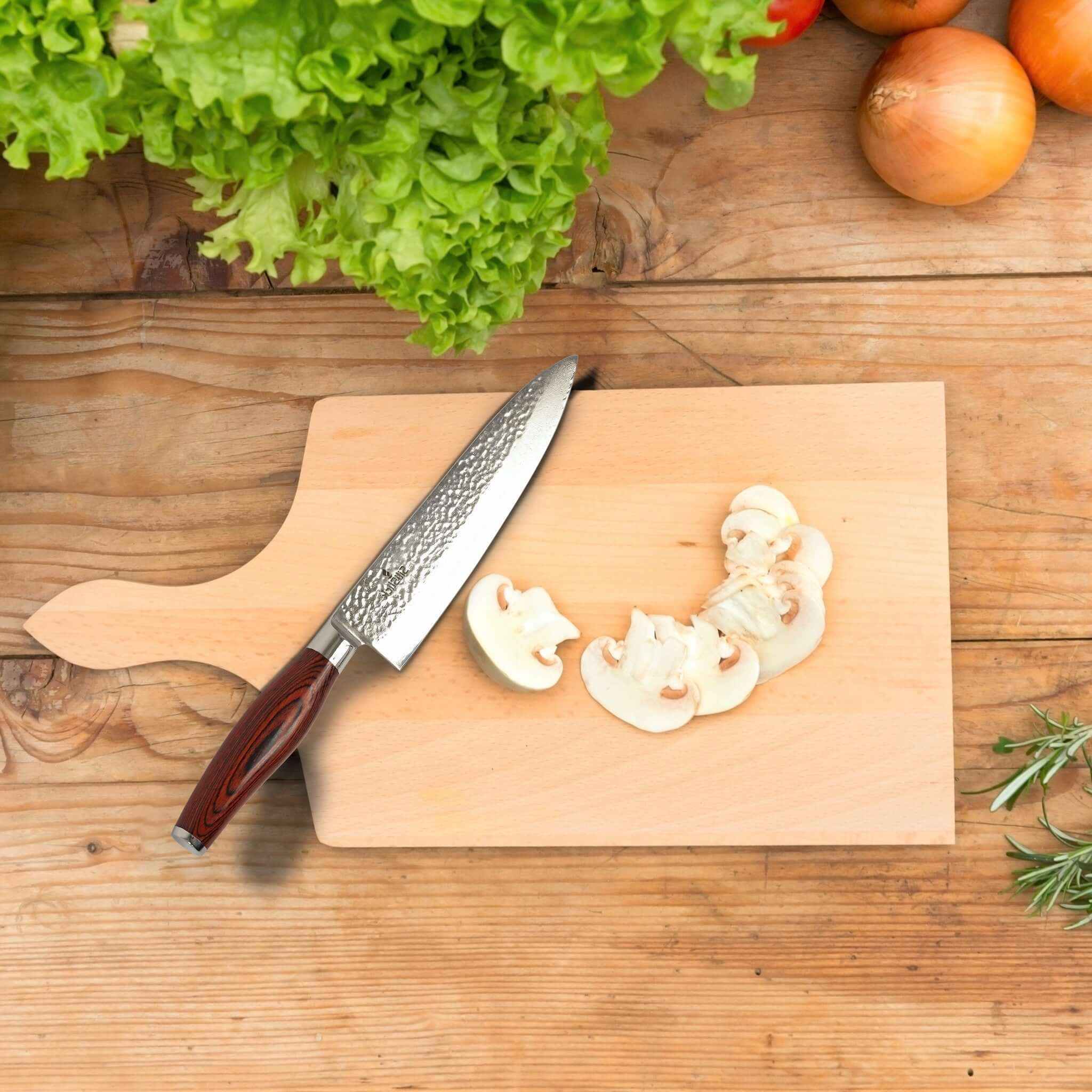 Damascus chef knife on a wooden board with sliced mushrooms and fresh produce