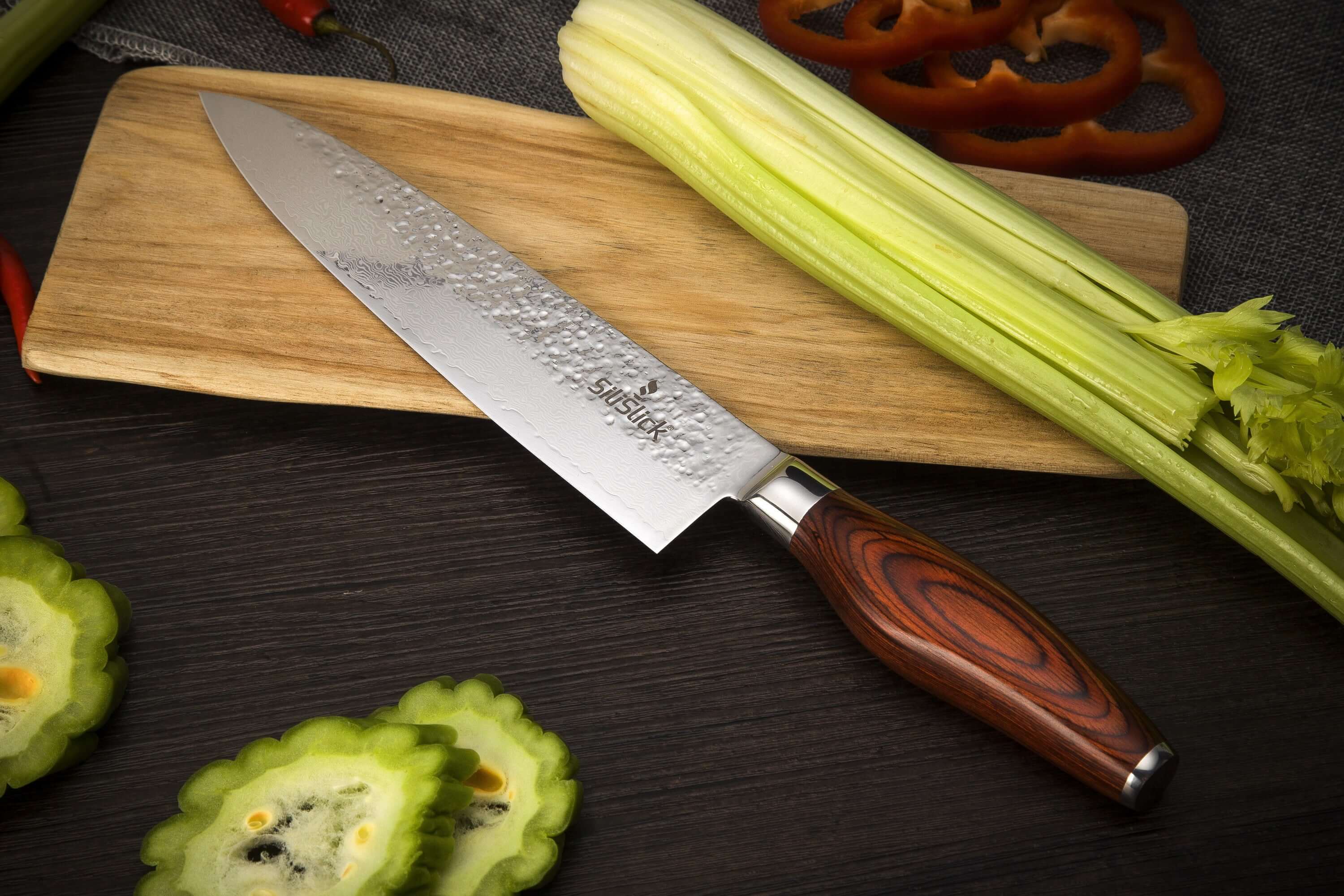 Damascus chef knife on a dark surface beside a cutting board and sliced vegetables