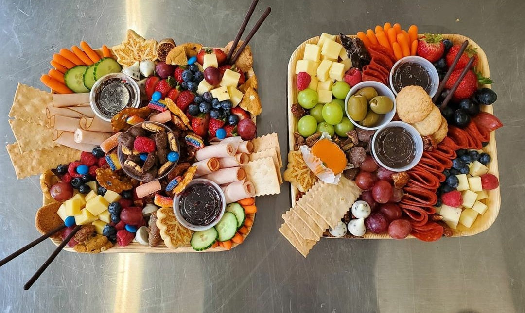 Two palm leaf trays laid out as grazing boards with meats, cheeses, fruit and crackers